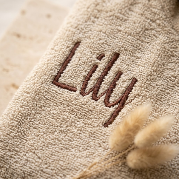 Een gedetailleerde macro-opname die scherpstelt op de geborduurde naam 'Lily' in donker garen op de zachte zandkleurige badstof van de badjas.