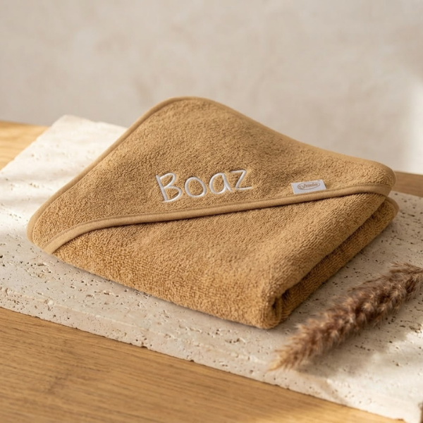 Een schuin bovenaanzicht van de opgevouwen camelkleurige badponcho met de witte geborduurde naam 'Boaz', gepresenteerd op travertijn en eikenhout.