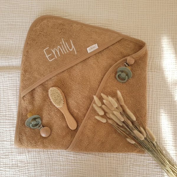 Een bovenaanzicht (flat-lay) compositie met de gepersonaliseerde 'Emily' badponcho, een houten babyborstel, een saliegroene speen en droogbloemen op een crème wafelstof ondergrond.