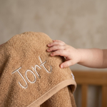 Een intieme lifestyle close-up waarin een babyhandje zachtjes de badstof van de camelkleurige badponcho met de geborduurde naam 'Jort' aanraakt.