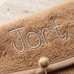 Een gedetailleerde macro-opname die scherpstelt op de taupekleurige geborduurde naam 'Jort' in een sierlijk lettertype op de zachte badstof.