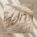 Close-up van een beige, zachte teddy- of badstof stof met een gedetailleerde borduring van een naam in een bijpassende lichtbruine draad.