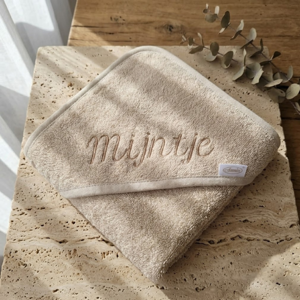 Beige/zandkleurige badstof wikkeldoek voor een baby met capuchon, geborduurd met de naam "Mijntje" in sierletters.
