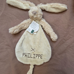 Beige pluche Happy Horse knuffeldoekje in de vorm van een konijn, met lange oren, een staartje en de naam 'PHILIPPE' erop geborduurd, liggend op een bruine ondergrond.